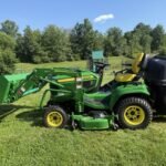 Green John Deere compact utility tractor with front-end loader and mowing deck, parked on a sunny grassy field.