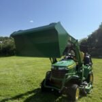 Green John Deere compact utility tractor with a raised front-end loader bucket, parked on a sunny grassy field.
