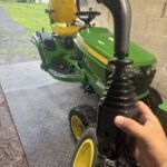 John Deere riding lawn mower with yellow seat and large rear wheels, parked on a metal ramp in a shed