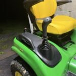 Green John Deere riding lawn tractor with a yellow seat and black joystick, parked on a metal surface near a cinder block wall.